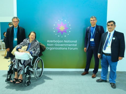 The image features four individuals standing in front of a backdrop displaying the logo of the "Azerbaijan National Non-Governmental Organizations Forum." From left to right: Viktor Sebek, Chairman of SSCC; Cecile Alvarez, SSCC Policy Advisory Board Member and UNESCO Artist for Peace, seated in a wheelchair; an unidentified individual; and Ramil Iskandarli, Chairman of Azerbaijan’s National NGO Forum. All participants are wearing event badges.