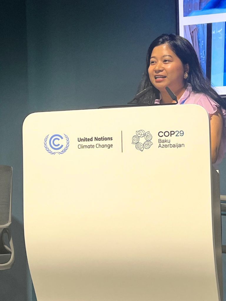 Dewy Sacayan speaking at a COP29 side event. She is standing behind a podium displaying the United Nations Climate Change and COP29 Baku Azerbaijan logos, addressing the audience on climate action.